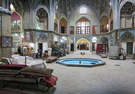 Are Persian Style Rugs a Good Investment? A Timeless Choice A view of the Aminoddole Carvansarai in the Bazaar of Kashan, Iran, featuring a central fountain and intricate architectural details, with various rugs and goods displayed.
