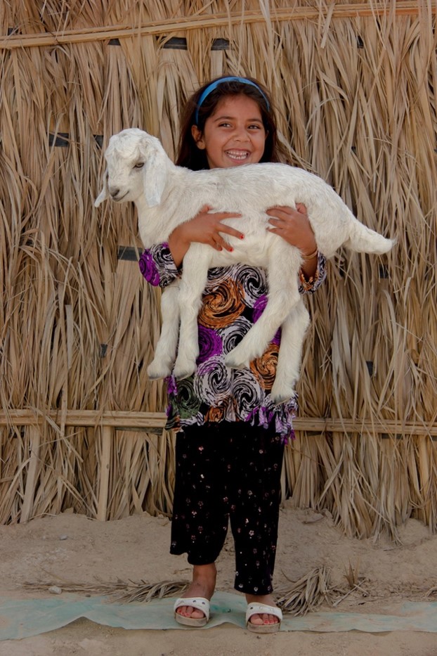 How Persian Rugs Are Made: Art and Tradition A young girl holds a small white lamb in front of a woven straw backdrop, showcasing the connection between pastoral life and the materials used in a Persian Style Rug. Photo by Mostafa Meraji
