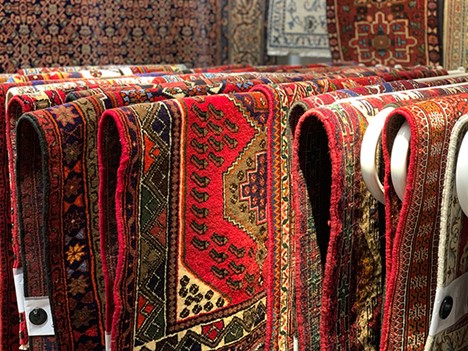 What Is an Oushak Rug? A Timeless Treasure A vibrant array of Persian rugs with intricate designs and rich colors are neatly displayed, likely in a market or shop. Photo by Ali Homayoun Nia
